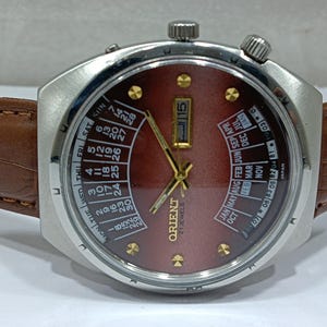 May include: A vintage Orient watch with a burgundy dial, gold hands, and a day-date calendar. The watch has a stainless steel case and a brown leather strap. The dial displays the day of the week and the month. The watch is marked "21 Jewels".