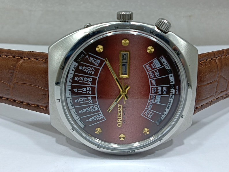 May include: A vintage Orient watch with a burgundy dial, gold hands, and a day-date calendar. The watch has a stainless steel case and a brown leather strap. The dial displays the day of the week and the month. The watch is marked "21 Jewels".