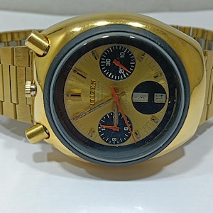 Peut inclure: Une montre chronographe automatique Citizen dorée avec un bracelet doré. Le cadran de la montre est doré avec des sous-cadrans noirs et des aiguilles rouges et orange. La montre a un affichage de la date et est de style vintage.
