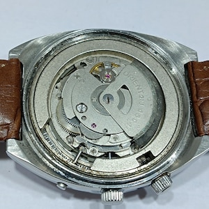 May include: Close-up of a vintage wristwatch with a silver-toned case and a brown leather strap. The watch's intricate internal mechanism is visible, featuring gears, springs, and the text "ORIENT 46944". The watch has a crown and a small button.