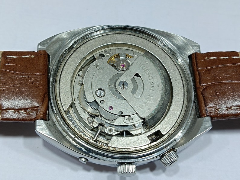 May include: Close-up of a vintage wristwatch with a silver-toned case and a brown leather strap. The watch's intricate internal mechanism is visible, featuring gears, springs, and the text "ORIENT 46944". The watch has a crown and a small button.
