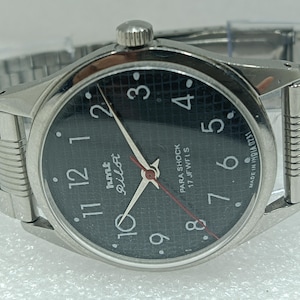 May include: Silver wristwatch with a black dial, white numerals, and hands. The watch face displays "hmt Pilot" and "PARA SHOCK 17 JEWELS". The watch has a silver metal strap.