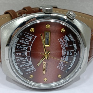 May include: A vintage Orient watch with a brown face, gold hands, and a stainless steel case. The watch features a brown leather band and a day and date display. The watch face displays the Orient logo and the words "21 Jewels".