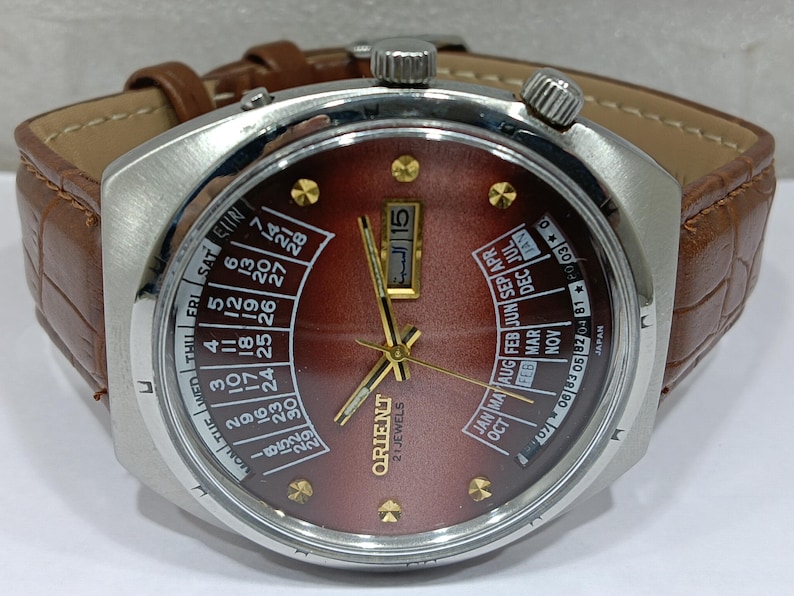 May include: A vintage Orient watch with a brown face, gold hands, and a stainless steel case. The watch features a brown leather band and a day and date display. The watch face displays the Orient logo and the words "21 Jewels".