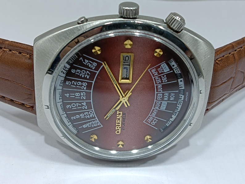 May include: A vintage Orient 21 Jewels wristwatch with a brown dial and calendar features. The watch has a stainless steel case, gold-tone hands and markers, and a brown leather strap. The dial displays the date and month, with Arabic numerals.