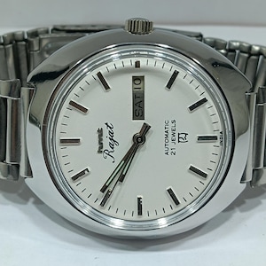 Op de afbeelding: Een zilverkleurig polshorloge met een witte wijzerplaat. De wijzerplaat toont de dag en datum, met het woord "Rajat" in cursief. Het horloge heeft een zilveren band en een ronde wijzerplaat. Het horloge leest "Automatic 21 Jewels" en "India".