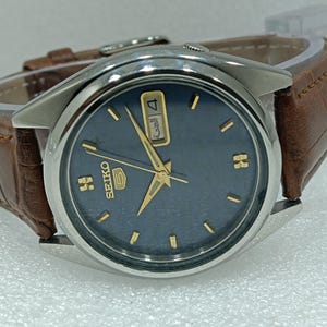 Puede incluir: Una taza de cerámica blanca con un asa negra y el texto "But First, Coffee" en negrita negra. La taza está sobre una superficie de madera.