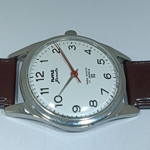 May include: A silver-toned wristwatch with a white face, black numerals, and a red second hand. The watch has a brown leather band and the text "HMT Janata" and "Para Shock 17 Jewels" on the face.