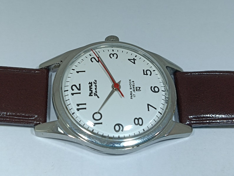 May include: A silver-toned wristwatch with a white face, black numerals, and a red second hand. The watch has a brown leather band and the text "HMT Janata" and "Para Shock 17 Jewels" on the face.