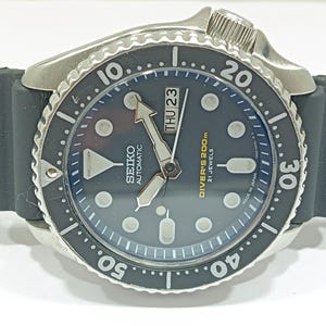 May include: A close-up of a Seiko automatic diver's watch with a black dial and bezel. The watch has a stainless steel case and a black rubber strap. The dial displays the day and date, with the text "DIVER'S 200m" and "21 JEWELS".