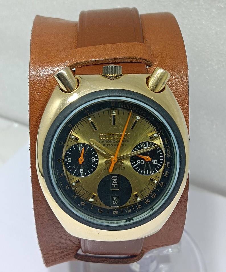 May include: A vintage gold-toned Citizen chronograph watch with a brown leather band. The watch face features black sub-dials, orange hands, and the date displayed at the bottom. The watch is attached to a wide brown leather cuff.
