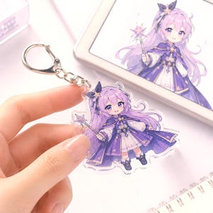 May include: A clear acrylic keychain featuring a chibi-style character with long purple hair, wearing a purple and white outfit with star accents, and holding a star-tipped wand. The character is depicted in a matching illustration on a tablet screen.