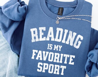 Könnte beinhalten: Ein marineblaues Sweatshirt mit dem Text "READING IS MY FAVORITE SPORT" in weißen Blockbuchstaben. Eine silberne Halskette mit einem kleinen Anhänger ist über dem Sweatshirt drapiert. Das Sweatshirt ist auf einer Jeans gefaltet.