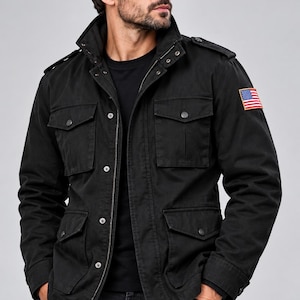 Op de afbeelding: Een zwart veldjack met een hoge kraag, ritssluiting aan de voorzijde met drukknopen, vier voorzakken met klep en epauletten op de schouders. Een kleine rechthoekige patch van de Amerikaanse vlag is op de linkermouw gestikt.