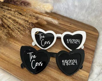 May include: Two pairs of heart-shaped sunglasses, one white and one black, with the text "The Coxs" and the date "11/9/2024" printed on the lenses. The sunglasses are displayed on a wooden surface.