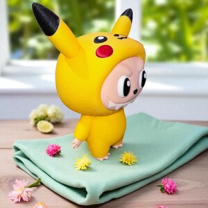 May include: A yellow Pikachu-themed figurine with black-tipped ears and red cheek circles. The figure is standing on a folded, light green cloth, surrounded by small pink and yellow flowers. The background is blurred, suggesting a natural setting. The figurine has a smiling face.