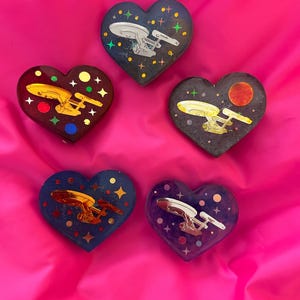 May include: Five heart-shaped resin ornaments featuring space-themed designs. Each piece displays a metallic starship silhouette against a dark, glittery background with colorful planet and star accents in shades of blue, purple, red, and black.