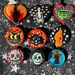 May include: A collection of Halloween-themed pins and brooches. The designs include a skeleton, a black cat, a skull, and a witch. The pins are in various shapes, including hearts and circles, and feature vibrant colors like orange, green, and black.