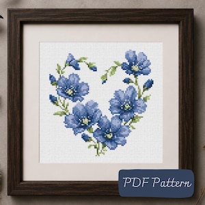 May include: A cross-stitch pattern featuring blue flowers arranged in a heart shape on a white grid background, framed in a dark wood square frame. A dark blue rectangular label in the bottom right corner displays the text PDF Pattern.