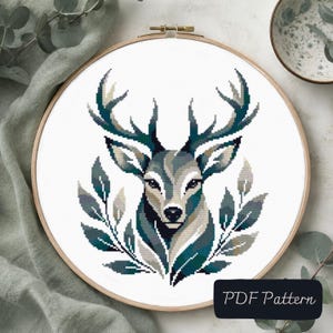 Peut inclure: Un projet de broderie au point de croix dans un tambour en bois circulaire représentant une tête de cerf stylisée avec de grands bois, dans des tons bleu sarcelle foncé, gris et blanc, entourée de motifs de feuilles. Étiquette PDF Pattern.