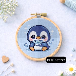 May include: Cross-stitch embroidery of a cute penguin holding a fish, set in a wooden hoop. The penguin is blue, white, and orange, with a light blue background. The text "PDF pattern" is at the bottom.