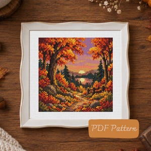 May include: A cross stitch pattern featuring a vibrant autumn landscape with orange and yellow trees framing a sunset over a lake. The design is displayed in a white frame and includes the text PDF Pattern on a brown rectangular label.
