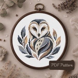 May include: A cross stitch pattern featuring a barn owl centred within a circular wooden embroidery hoop. The owl is rendered in shades of grey, tan, and white, surrounded by stylised leaf motifs in matching tones. The text PDF Pattern is visible.