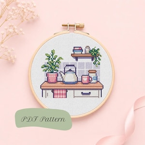 May include: A cross stitch pattern in a wooden embroidery hoop featuring a kitchen scene with a potted plant, a kettle, a mug, and a jar on a counter, with a shelf above holding books and a trailing plant, labelled with the text PDF Pattern.