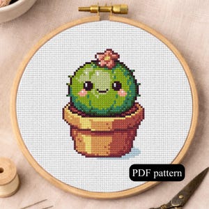 Puede incluir: Patrón de punto de cruz de un cactus verde sonriente en una maceta marrón, enmarcado en un bastidor de bordado de madera. El cactus tiene una pequeña flor rosa. El texto "PDF pattern" está en un recuadro negro.
