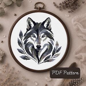 May include: A cross-stitch embroidery project featuring a stylized wolf head in shades of gray, black, and white, framed in a circular wooden hoop. The wolf is surrounded by leaf-like patterns. A dark rectangular label reads PDF Pattern.