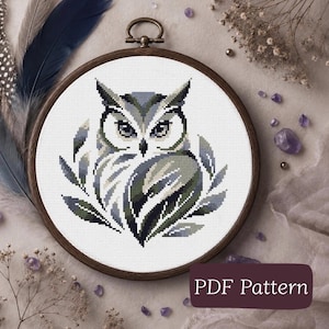 May include: A cross-stitch embroidery project featuring a stylized owl in shades of gray, green, and black, centered within a circular wooden hoop. The design is stitched on white fabric and includes the text PDF Pattern in a dark purple box.