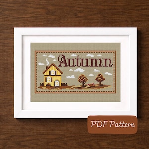 Könnte beinhalten: Ein Kreuzstich-Stickmuster in einem weißen Rahmen mit einem gelben Haus, zwei Bäumen und weißen Wolken auf beigem Stoffhintergrund. Das Wort Autumn ist in dunkelbraunen Buchstaben über dem Haus gestickt. Ein braunes Etikett zeigt PDF Pattern.