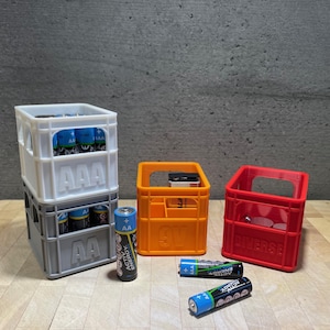 Könnte beinhalten: Drei Miniatur-Plastikkisten, eine weiße, eine graue und eine orange, mit den Worten "AAA", "AA" und "GT" bedruckt. Die Kisten sind mit AA- und AAA-Batterien gefüllt. Auf der Holzoberfläche liegen auch mehrere Batterien.