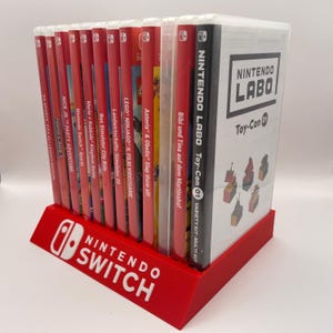 Può includere: Un supporto in plastica rosso con le parole "Nintendo Switch" in lettere bianche. Il supporto contiene una varietà di cartucce di gioco per Nintendo Switch, tra cui "EA Sports FIFA 22 Legacy Edition", "Minelart", "Nick Jr. Party Adventure", "Nintendo Switch Sports", "Mario + Rabbids Kingdom Battle", "Bus Simulator City Ride", "Farming Simulator 20", "LEGO Ninjago - The Film Videogame", "Asterix & Obelix Slap them all!", "Bibi und Tina auf dem Martinshof" e "Nintendo Labo Toy-Con 01 Variety Kit Multi-Kit".