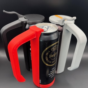 May include: Three 3D printed can holders in red, gray, and black. The holders are designed to fit around a standard aluminum can and have a handle for easy carrying. The can in the red holder has a black label with gold lettering that reads "Warsteiner, Eine Königin unter den Bieren".