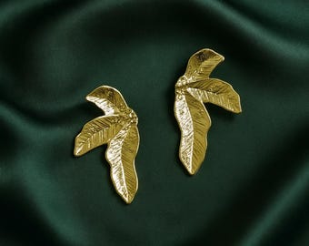 May include: A pair of gold-colored leaf-shaped earrings. The earrings are detailed with veining and are set against a dark green satin background. The leaves are facing opposite directions.