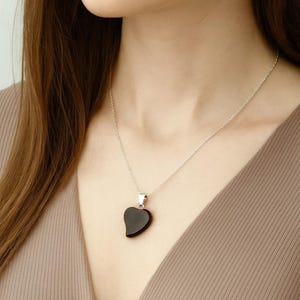 Può includere: Una collana d'argento con un ciondolo nero a forma di cuore. Il ciondolo è liscio e lucido, sospeso a una sottile catena d'argento. La collana è indossata su un top dai toni neutri, creando un look semplice ed elegante.