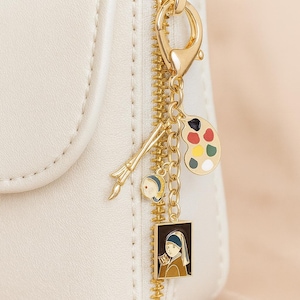 May include: Gold charm cluster featuring art-related pendants: a paintbrush, a palette, and a miniature painting of a woman. The charms are attached to a white purse with a gold zip via a gold clasp.