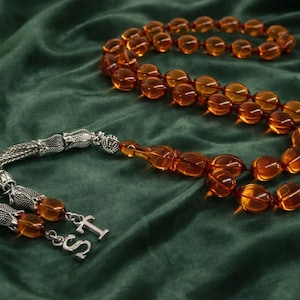 Puede incluir: Un collar de cuentas de oración de color ámbar con detalles plateados, sobre una superficie de terciopelo verde oscuro. Las cuentas ovaladas están ensartadas, con una borla plateada decorativa y un colgante plateado.