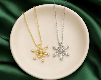 以下が含まれることがあります： 白いお皿の上に、金色のものと銀色のものの2つの雪の結晶ペンダントネックレスが置かれています。緑色の背景です。各ネックレスは繊細なチェーンと、小さな輝くクリスタルで飾られた雪の結晶ペンダントが特徴です。