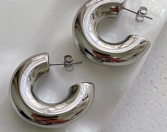 May include: A pair of shiny, silver-colored hoop earrings. The earrings are thick and rounded, with a smooth, reflective surface. They are displayed on a white surface, showcasing their polished appearance. The earrings have a post and butterfly backing.