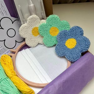 Könnte beinhalten: Ein Punch-Needle-Set mit einem hölzernen Stickrahmen, weißem Stoff, Garnbündeln in Minzgrün und Gelb, einem Punch-Needle-Werkzeug sowie drei fertigen blumenförmigen Untersetzern in Weiß, Minzgrün und Blau mit gelben Mittelpunkten.
