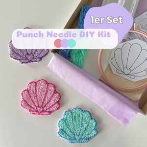 Könnte beinhalten: Ein Punch-Needle-DIY-Set mit einem hölzernen Stickrahmen, Stoff, blauem und grünem Garn sowie einer muschelförmigen Vorlage. Drei fertige muschelförmige Punch-Needle-Untersetzer in Lila, Pink und Grün liegen neben der Box.