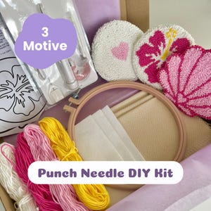 Könnte beinhalten: Ein Punch-Needle-DIY-Set mit einem hölzernen Stickrahmen, beigem Stoff, weißem Filz, einem Punch-Needle-Werkzeug und Garnsträngen in Weiß, Pink, Magenta und Gelb. Drei fertige runde Punch-Needle-Untersetzer zeigen Herz-, Hibiskus- und Muschel-Motive.