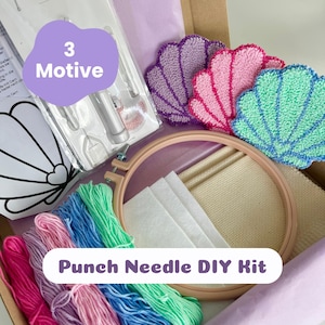 Könnte beinhalten: Ein Punch-Needle-DIY-Set mit einem hölzernen Stickrahmen, beigem Stoff, weißen Filzstücken, einem verpackten Punch-Needle-Werkzeug und mehreren Garnsträngen in Pink, Lila, Blau und Grün. Drei fertige Untersetzer in Form von Muscheln sind enthalten.