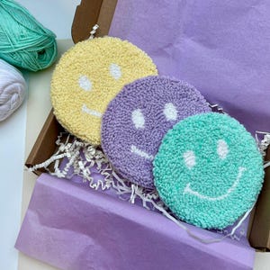 Könnte beinhalten: Ein Set aus drei runden, getufteten Untersetzern in einer Box. Jeder Untersetzer hat ein Smiley-Gesicht-Design mit weißen Augen und einem gestickten Lächeln. Die Untersetzer sind gelb, lila und türkis. Die Box ist braun mit lila Seidenpapier und weißem Papierfüllmaterial.