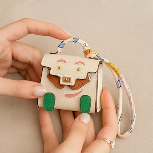 Op de afbeelding: Een kleine, crèmekleurige handtas met een cartoongezichtontwerp. De tas heeft bruine accenten, groene voetjes en een kleurrijk patroonhandvat. De tas wordt door twee handen vastgehouden.