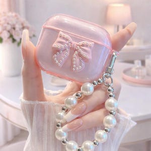 May include: A pink, glittery AirPod case with a 3D bow adorned with small pearls. The case has a silver clasp and is attached to a pearl bracelet. The overall aesthetic is feminine and elegant.