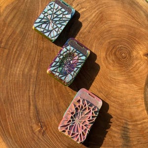 Könnte beinhalten: Drei schillernde rechteckige Etuis mit geometrischen Designs. Die Etuis sind in Blau-, Grün-, Lila- und Rosatönen gehalten und weisen ein Sternmuster auf. Sie sind auf einer Holzoberfläche angeordnet.