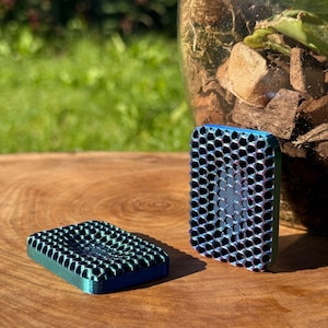 May include: Two small, rectangular, 3D-printed objects with a honeycomb pattern. One is lying flat, the other is standing upright. The objects have a gradient color scheme, transitioning from teal to blue. They are placed on a wooden surface.
